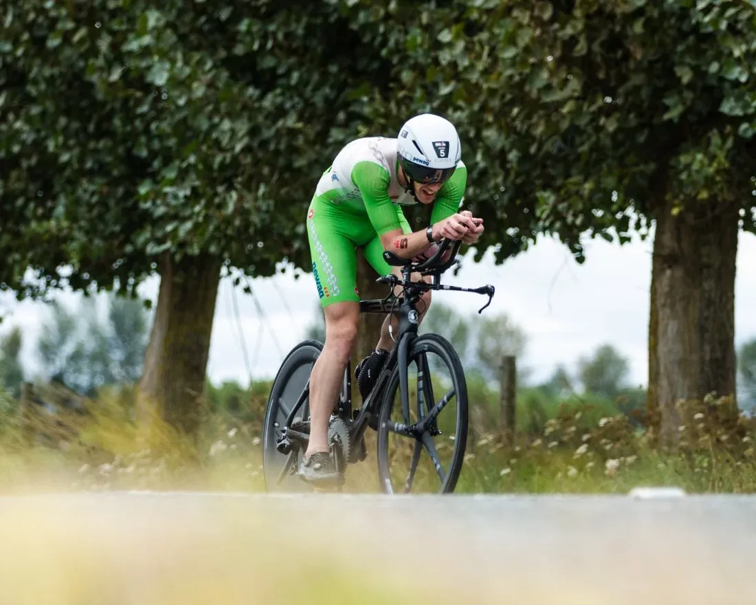 A triathlete in an aero tuck on a time trial bike, riding a tree-lined country road
